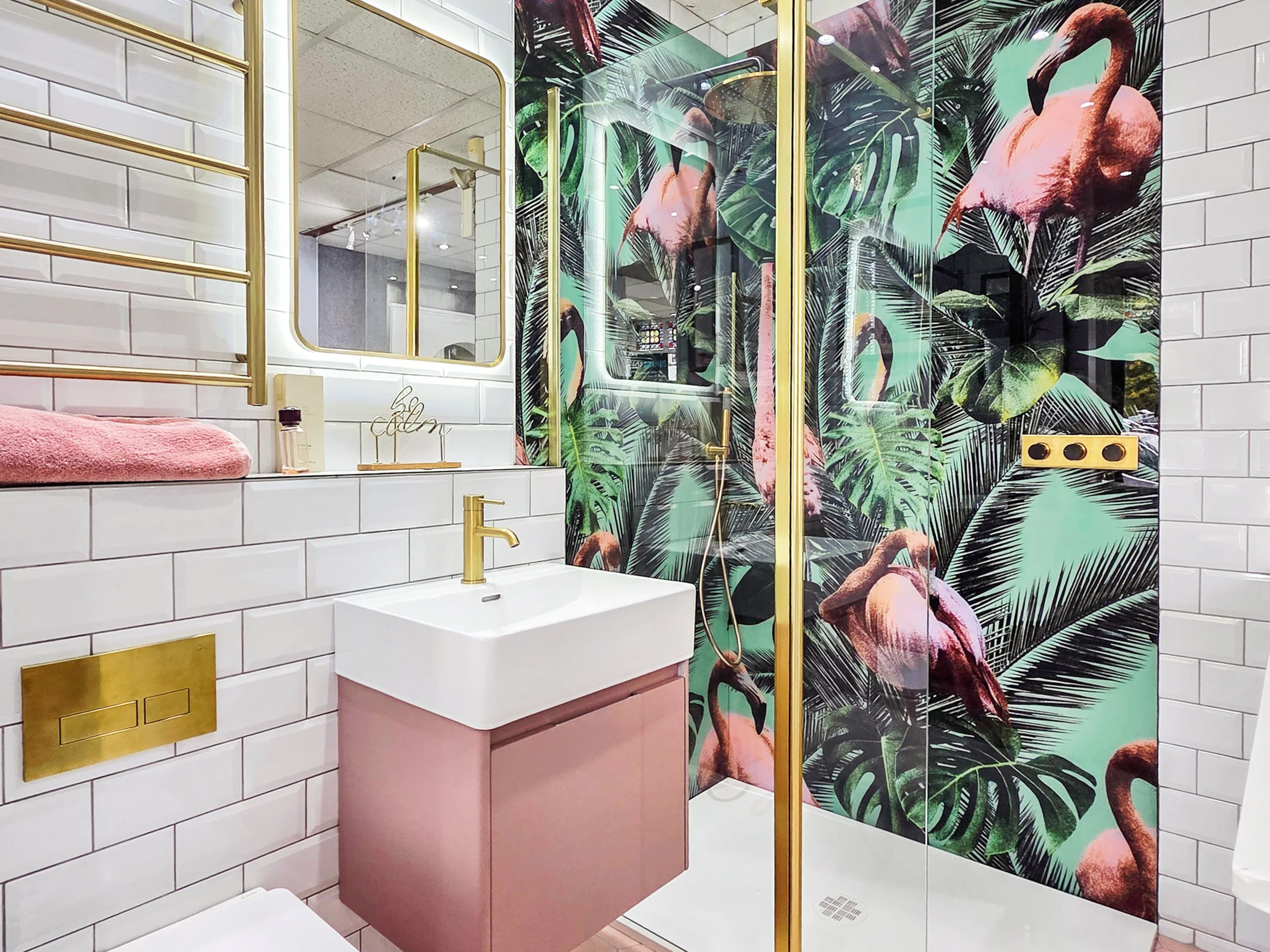 Designer bathroom featuring a pink vanity unit, brass fittings, illuminated mirror, tropical flamingo feature wall, white subway tiles, and a luxury walk-in shower in the Love Your Bathroom showroom.
